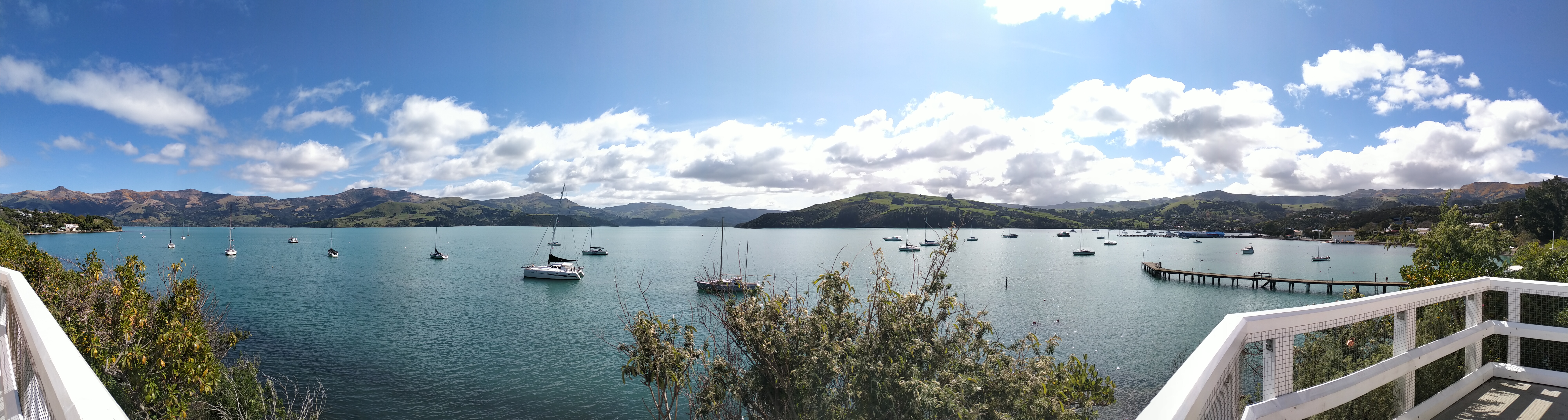 A panoramic view of Akaroa Harbour on a sunny day, with the Port Hills in the background and a number of boats sitting on the water