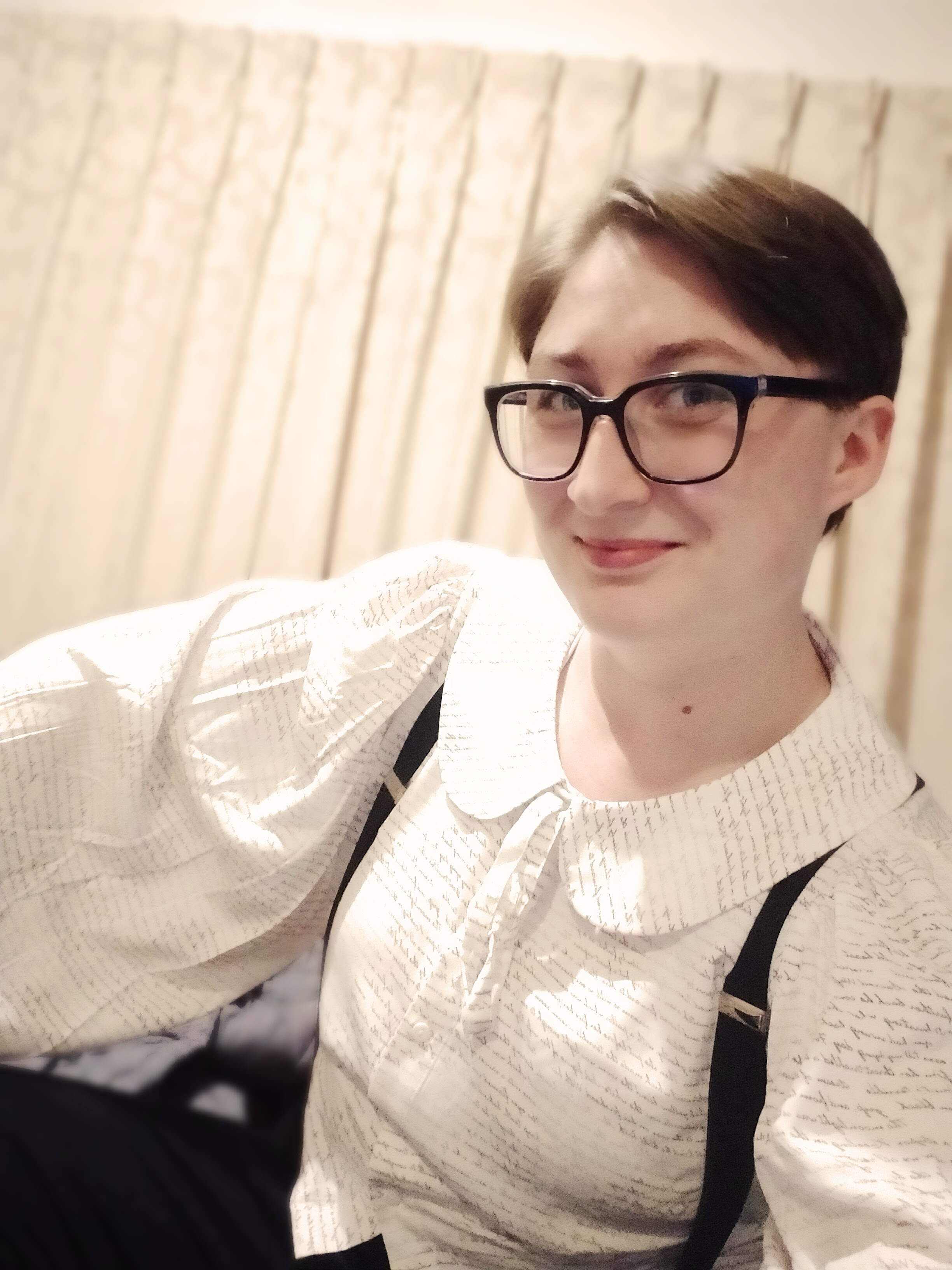 An androgynous person with short brown hair and fair skin smiling at the camera; they are wearing thick-rimmed glasses, a white, balloon-sleeved shirt, black suspenders, and black trousers.
